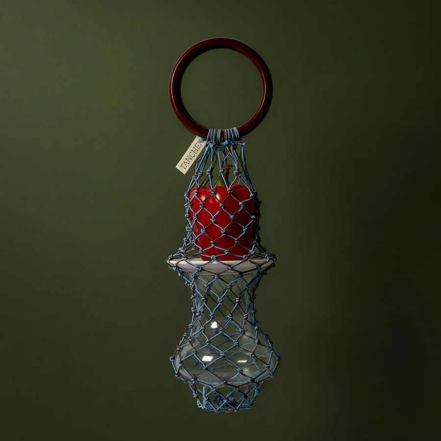 Styled image of the 00 Bag in Blue Brown by Tanchen, featuring the bag holding a glass bottle and apple against a dark green background.