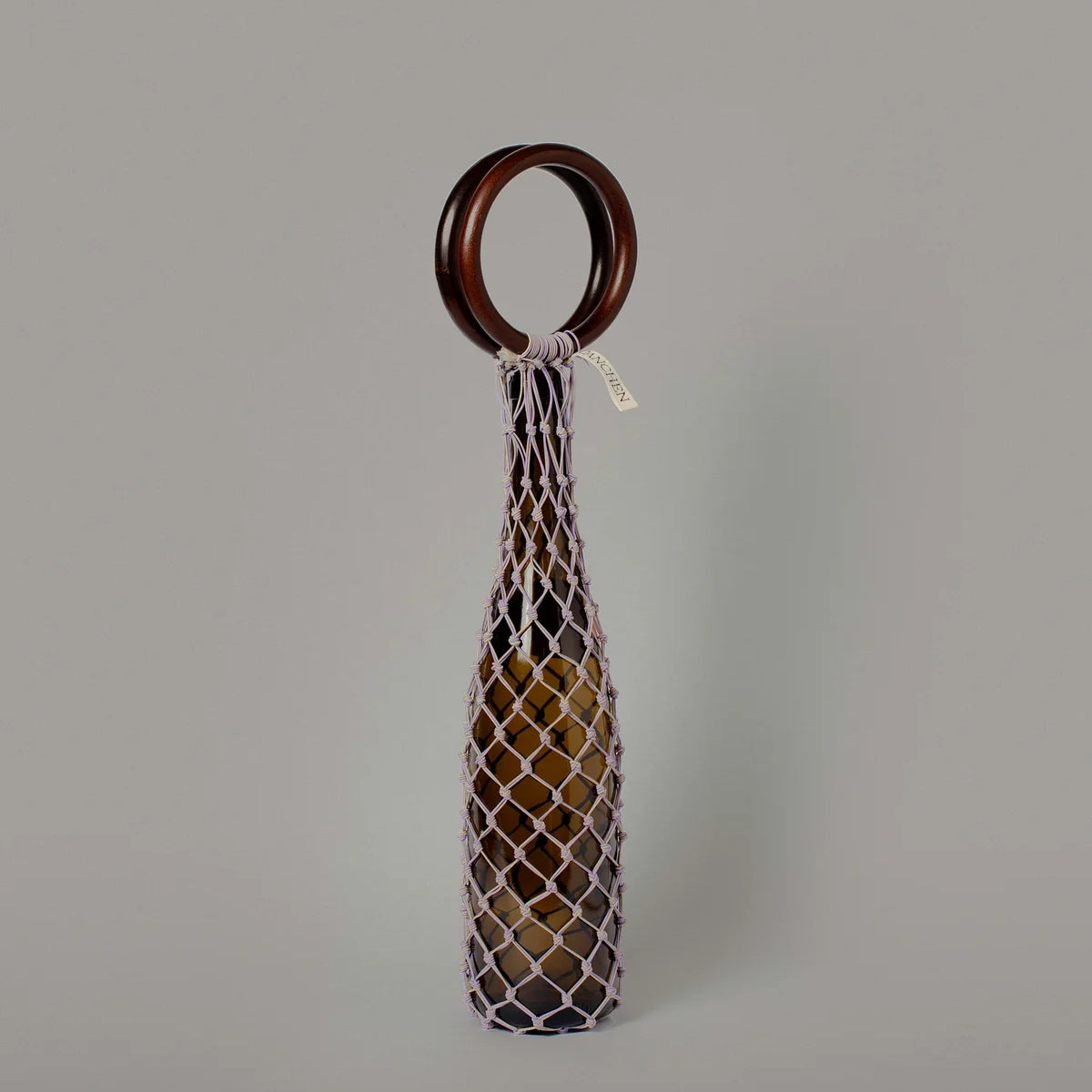 Tanchen 0/0 Bag in Lilac Sand displayed with a bottle, featuring a wooden ring and woven net structure.
