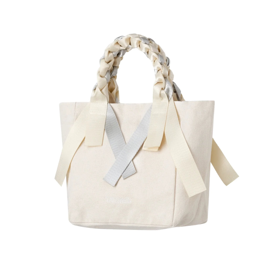 Side angle of the Tanchen Picnic Bag Mini Soda in cream and silver, highlighting the intricate braided handle and ribbon detailing on the body. The cream canvas and silver ribbon create a minimalistic yet elegant look.