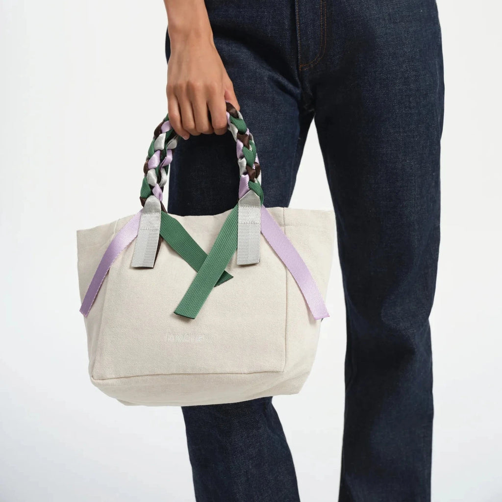 Tanchen Picnic Bag Mini Spritz held by a model wearing dark denim jeans, showcasing its cream cotton canvas with braided multi-coloured nylon ribbon handles.
